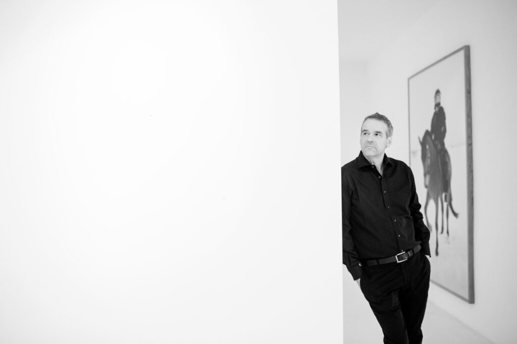 echtes Charakterportrait von dem Galeristen Stephan Stumpf in München. Er lehnt an der Wand in seiner Galerie, im Hintergrund ist das Kunstwerk Reiterstandbild von ANNA BARANOWSKI zu sehen