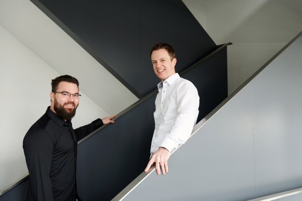 Cooles Corporatefoto in München. Zwie Mitarbeitende unterhalten sich auf der Treppe des Bürogebäudes und schauen freundlich in die Kamera.