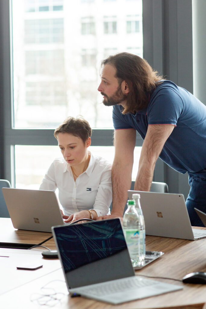 Zwei Mitarbeitende einer IT Firma am Laptop im Besprechungsraum, im Vordergrund sind weitere Laptops zu sehen.