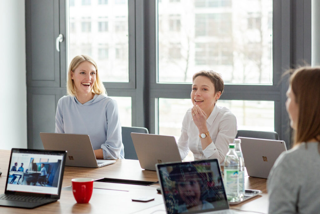 Lockere Corporatefotos München aus dem Besprechungsraum. Zwei Mitarbeiterinnen sind im Fokus, sie sitzen vor ihren Laptops und sind im Austausch mit anderen. Die Stimmung ist fröhlich.