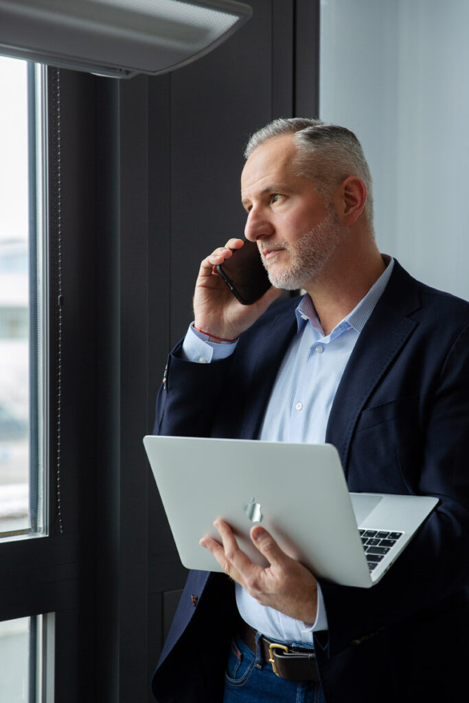 Professionelles Corporatefoto von Mitarbeiter in München mit Laptop in der Hand beim Telefonieren.