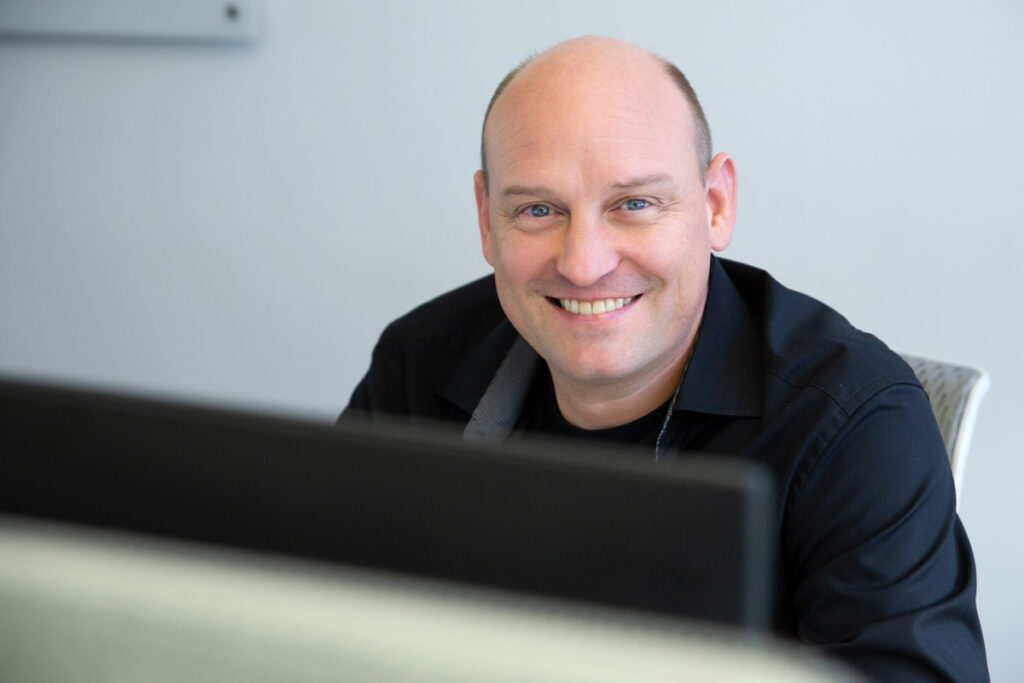 Starkes Corporatefoto von Mitarbeiter einer Firma in München, der von seiner Arbeit am PC mit einem sympathischen Lächeln aufblickt