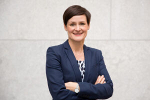 Authentisches Businessportrait von Frau im Business-Outfit mit verschränkten Armen vor Steinfassade im natürlichen Licht.