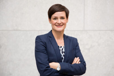 Authentisches Businessportrait von Frau im Business-Outfit mit verschränkten Armen vor Steinfassade im natürlichen Licht.