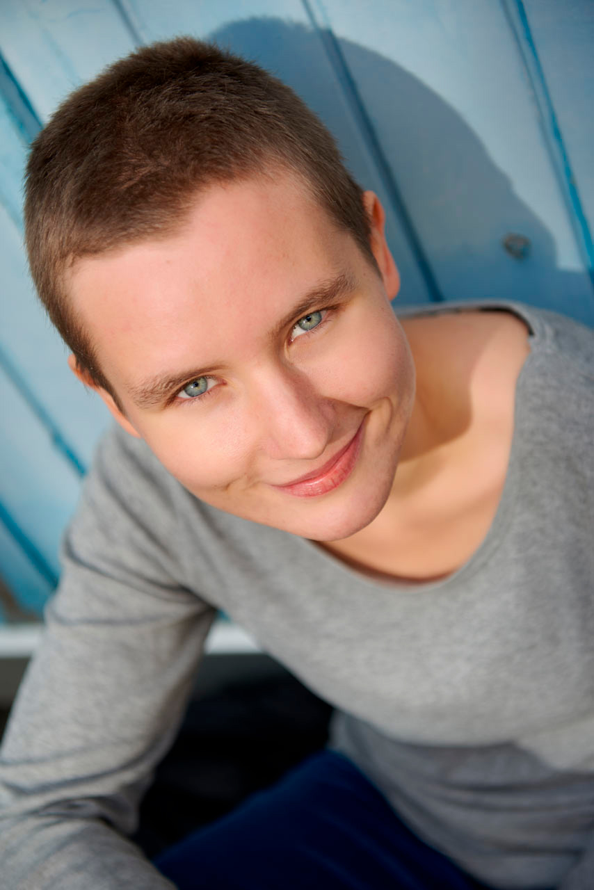 Sympathisches Autorenporträt: Close Up von junger Autorin im Innenhof vor blauer blauer Holzwand bei natürlichem Licht.