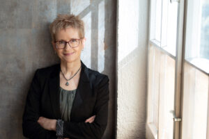 Modernes Businessportrait: Frau lehnt mit verschränkten Armen an einer Steinwand, das Licht fällt duch das Fenmster auf Sie und den Hintergrund.
