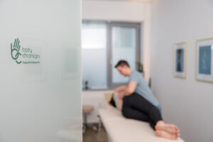 Corporate Fotografie von Osteopathie in Germering: im Vordergrund iszt die Tür mit dem Firmenlogo zu sehen, im Hintergrund in der Unschärfe behandlet der Therapeut eine Patientin am Rücken.
