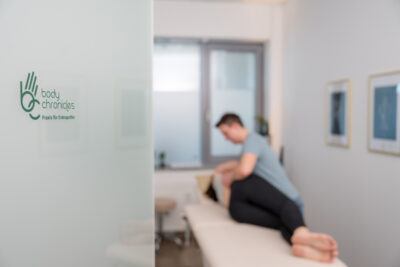 Corporate Fotografie von Osteopathie in Germering: im Vordergrund iszt die Tür mit dem Firmenlogo zu sehen, im Hintergrund in der Unschärfe behandlet der Therapeut eine Patientin am Rücken.