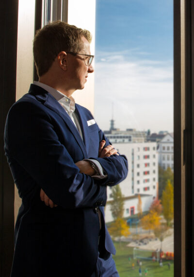Professionelles Corporate Portrait: Geschäftsführer lehnt mit verschränkten Armen an der Wand eines moderenen Büros in München und blickt aus dem Fesnter. Im Hintergund ist unscharf die Silhouette der Stadt zu sehen.