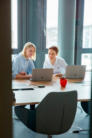 Auf dem Employer Branding Foto für eine IT Firma in München ist ein zweier Team im Gespräch mit Laptops, in einem Raum mit viel Glas.
