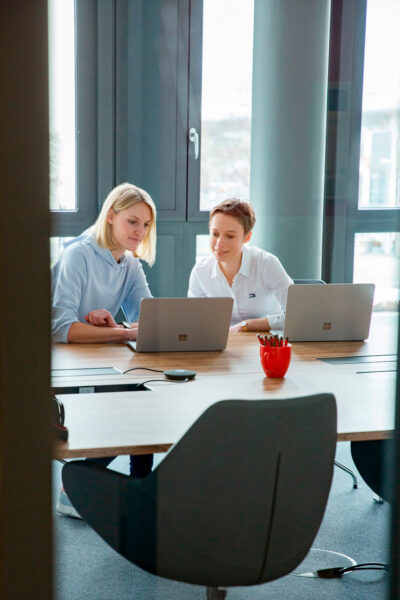 Auf dem Employer Branding Foto für eine IT Firma in München ist ein zweier Team im Gespräch mit Laptops, in einem Raum mit viel Glas.