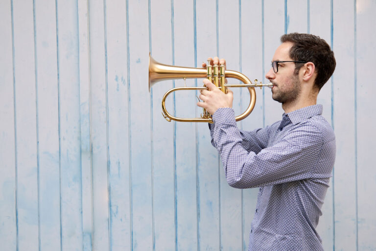 Kuenstleprotrait: Musiker spilet Trompete vor blauer Holzwand mit natürlichem Licht fotografiert.