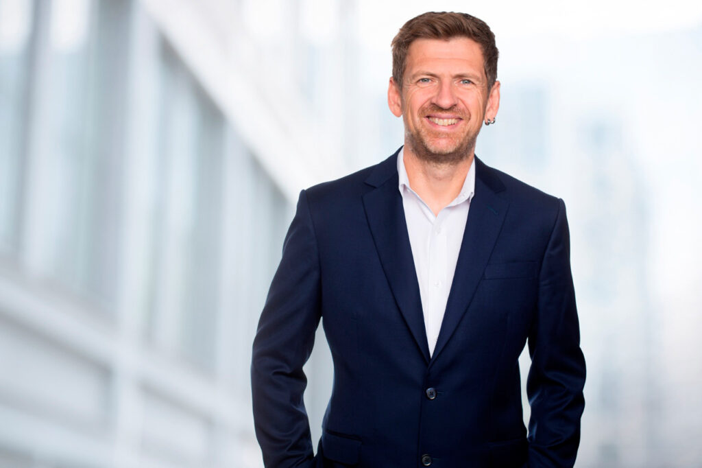 Modernes Bewerbungsbild eines Managers vor weichgezeichneter moderner Architektut in München mit Fokus auf Souveränität,