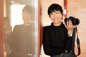 Portraitfoto von Künstlerin mit Spiegelung. Sie hält lächelnd eine Kamera in der Hand.