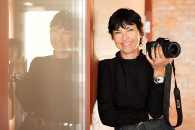 Portraitfoto von Künstlerin mit Spiegelung. Sie hält lächelnd eine Kamera in der Hand.