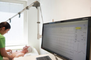 Praxisfotografie in Münchenr Arztpraxis. NAhaufnahme von Bildschrim mit EKG-Kurve, im Hintergrund unscharf Patient und Arzthelferin.