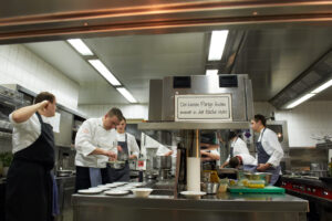 Unternehmensfotografie Gastronomie: Sinnspruch im Fokus, im Hintergrund verschwommenes, professionelles Küchenteam bei der Arbeit.