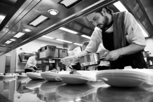 Unternehmensfotografie in schwarz-weiss mit spannender Perspektive: ein Koch richtet das Essen auf den Tellern im Berggasthof Aschbach an.