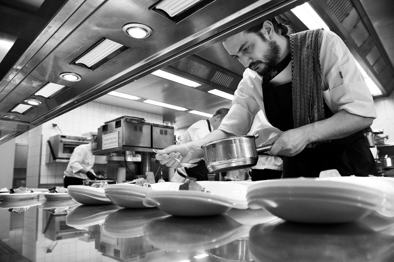 Unternehmensfotografie in schwarz-weiss mit spannender Perspektive: ein Koch richtet das Essen auf den Tellern im Berggasthof Aschbach an.